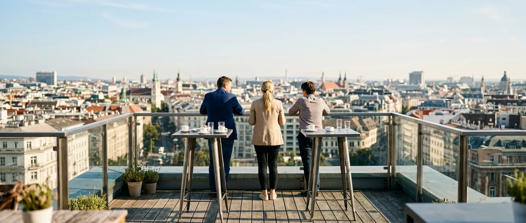 Drei Personen stehen an Stehtischen auf einer Dachterrasse und blicken über die Dächer einer Stadt.