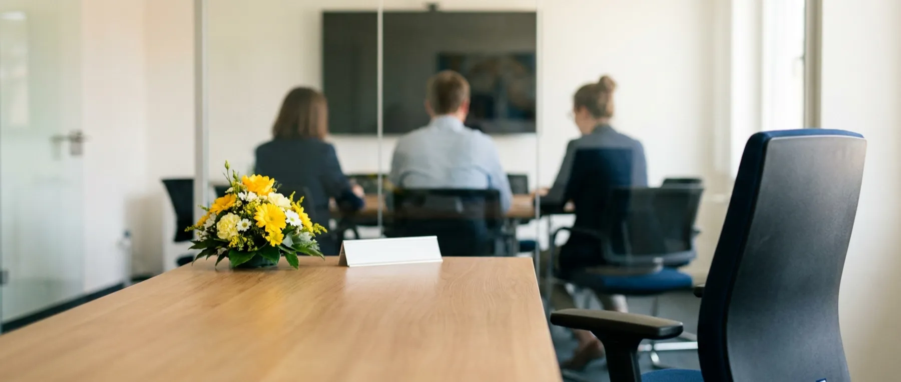 Ein Besprechungstisch mit Blumengesteck und umgeklapptem Namensschild, dahinter unscharf drei Personen im Gespräch.